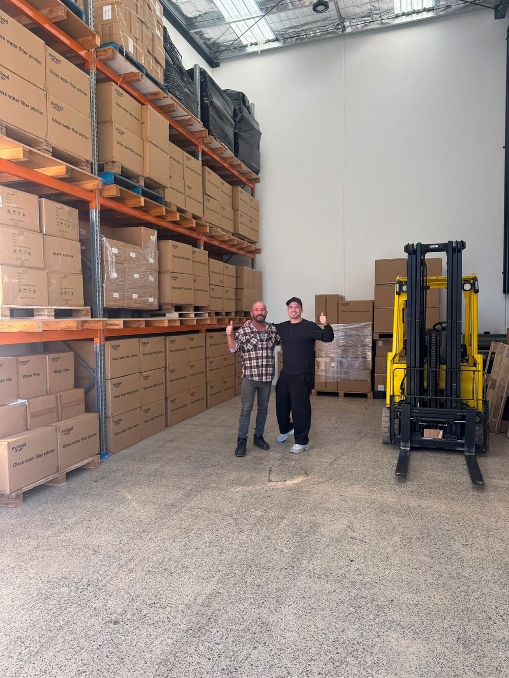 Two team members give a thumbs up beside full pallet racking and a yellow forklift.
